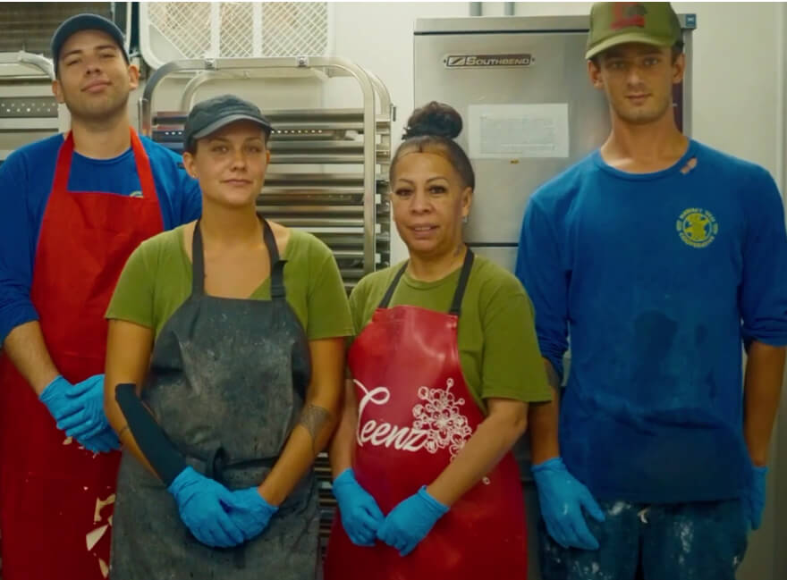 A group of staff workers in an industrial kitchen. The are wearing their work clothes, including colorful aprons, and look as if they are in the middle of their work day. They are wearing gloves which indiciate that they are working with food or food processing activities. A man on the right is wearing a blue shirt with a Hawaii Ulu Co-op logo which suggests that they are working at a Hawaii Ulu Co-op manufacturing facility.